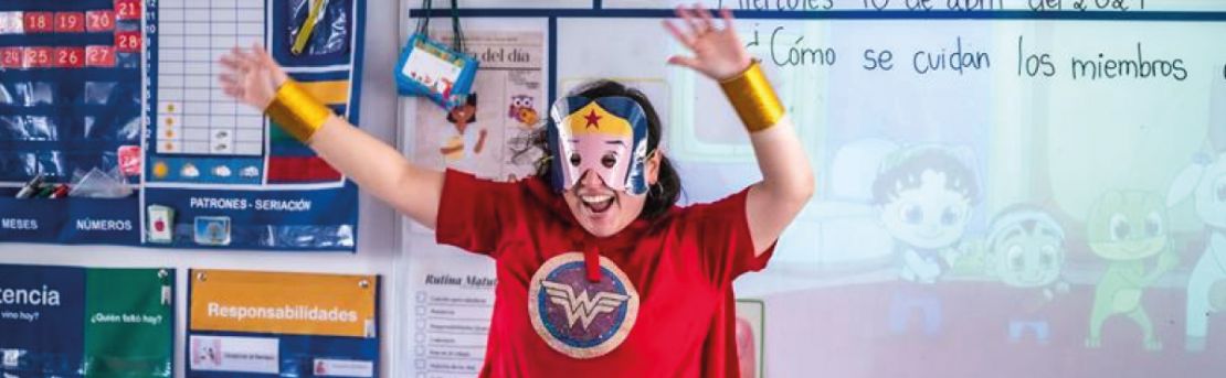 Profesora disfrazada de heroína alzando los brazos frente a una pizarra en un salón de clases.